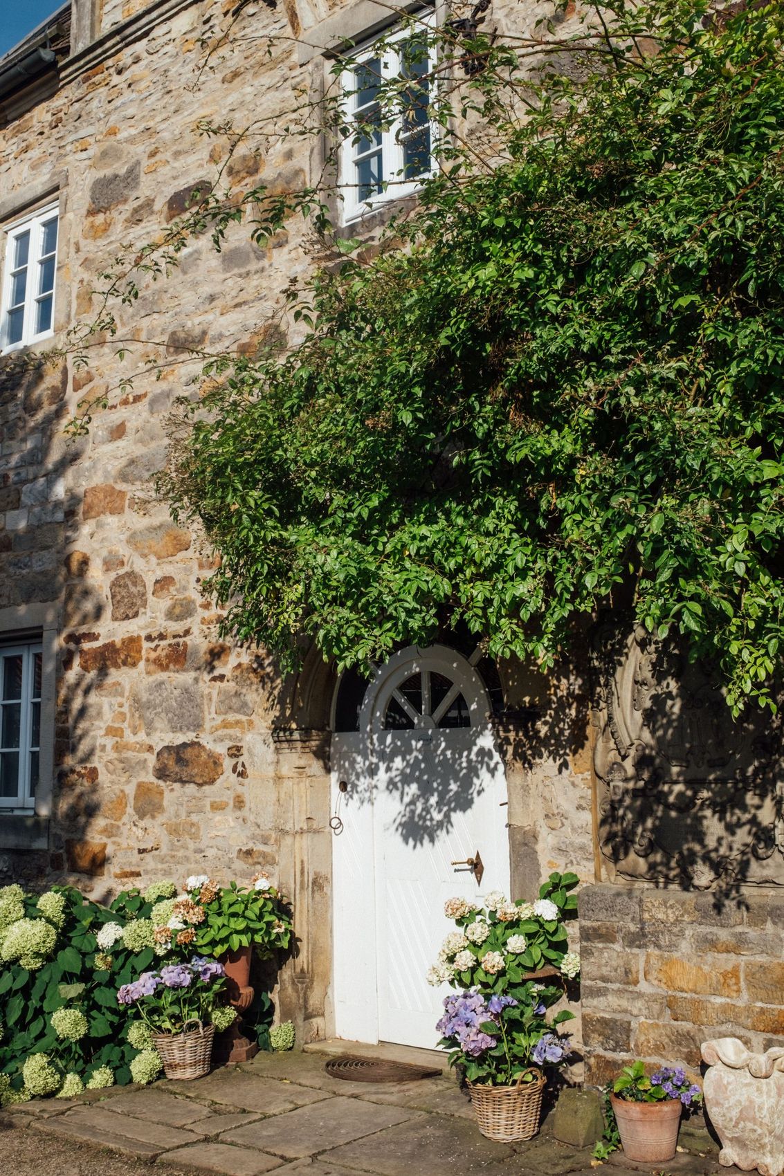 Honey-coloured stone cottage with a white painted door and flowers beside the step